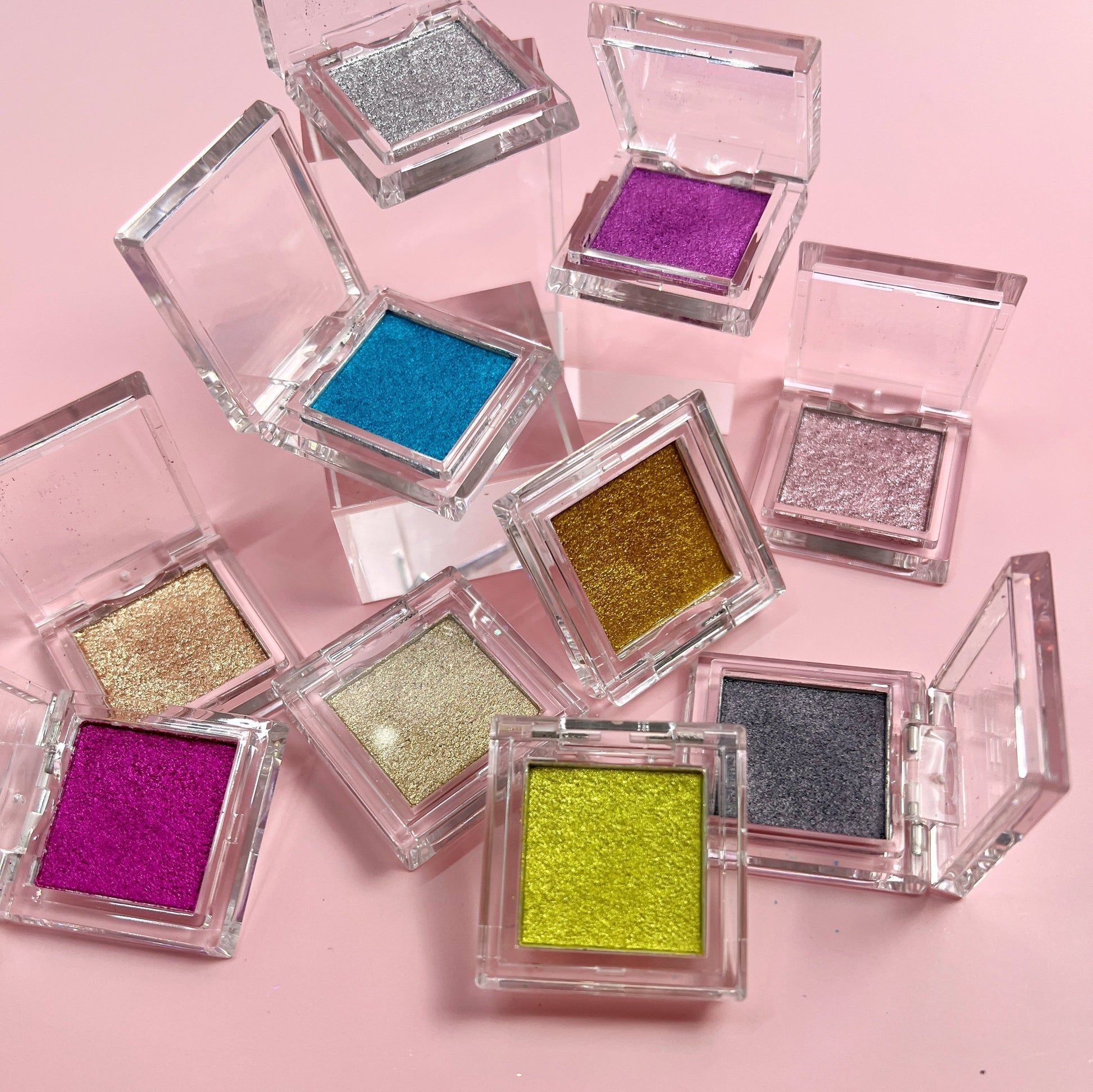 Clear compacts with multicolor metallic pressed powder scattered on pink background. 