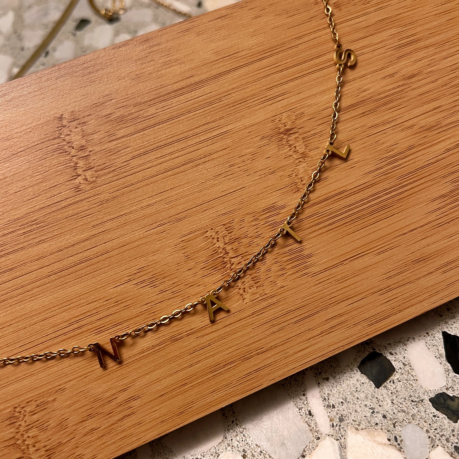 Gold chain necklace with letter charms on wood and terrazzo background. 